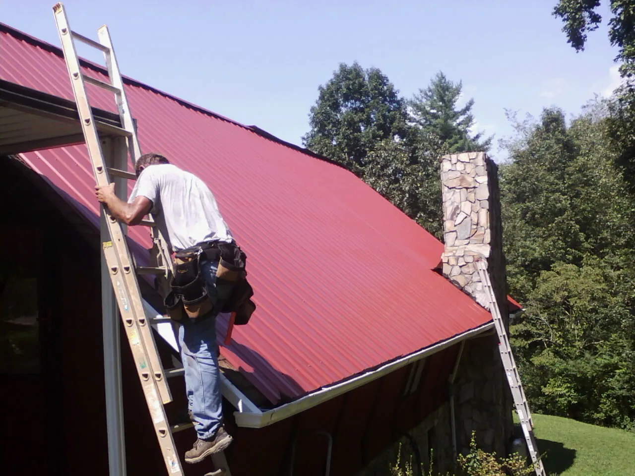 Roofer on a ladder beside a red metal roof and stone chimney — Soffit & Fascia Repair by Roofing Brothers in Coal City